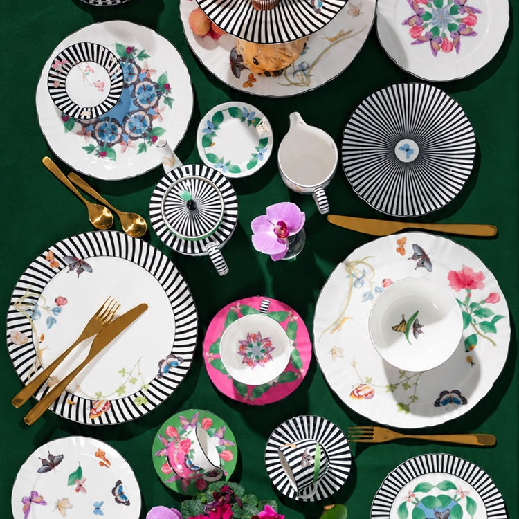 Koyu yeşil örtü üzerinde Kaleidoscope koleksiyonunun siyah-beyaz çizgili, çiçek ve kelebek desenli tabakları, fincanları ve demliği.